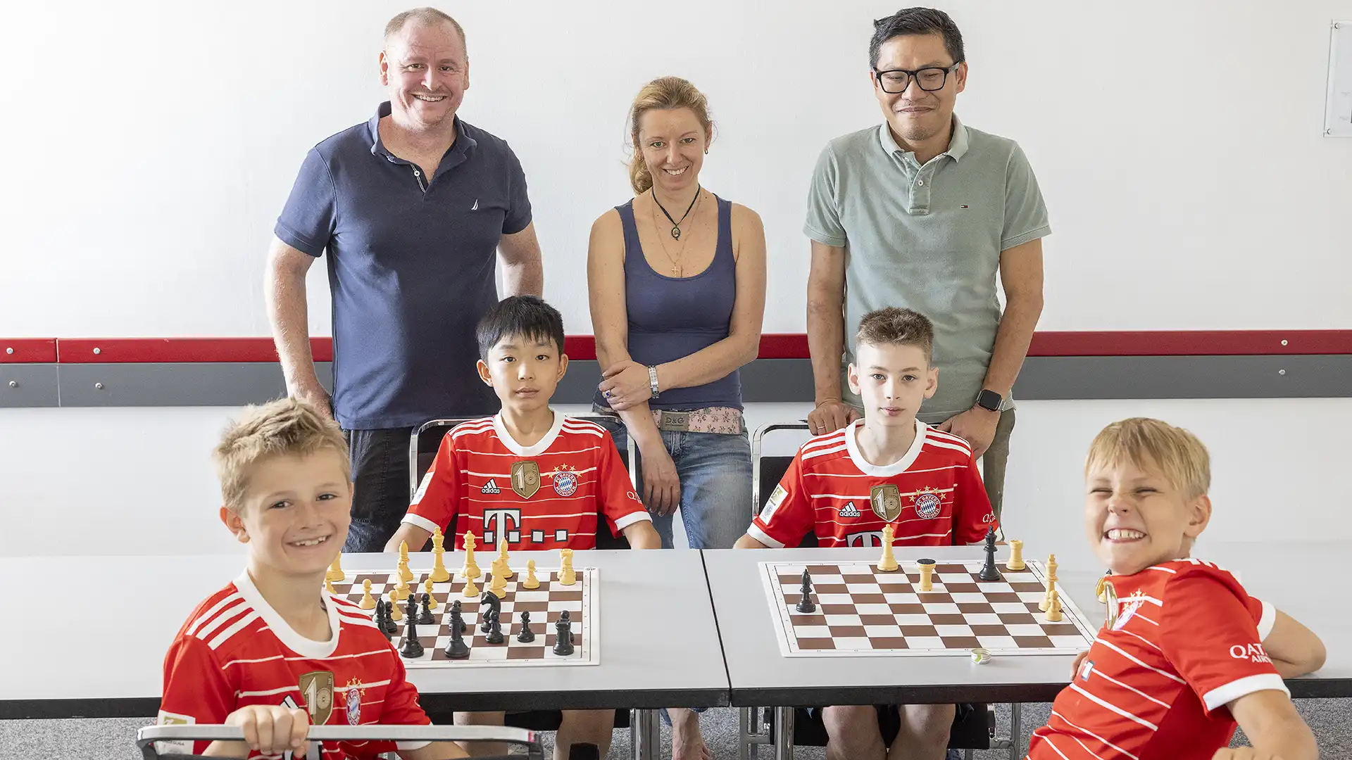 Farbfoto: Die junge Schach-Mannschaft des FC Bayern München mit 4 Kindern sitz an einem Tisch mit 2 Schachbrettern. Dahintrer stehen drei Eltern, die in Düsseldorf dabei waren. Alle gucken zum Betrachter.