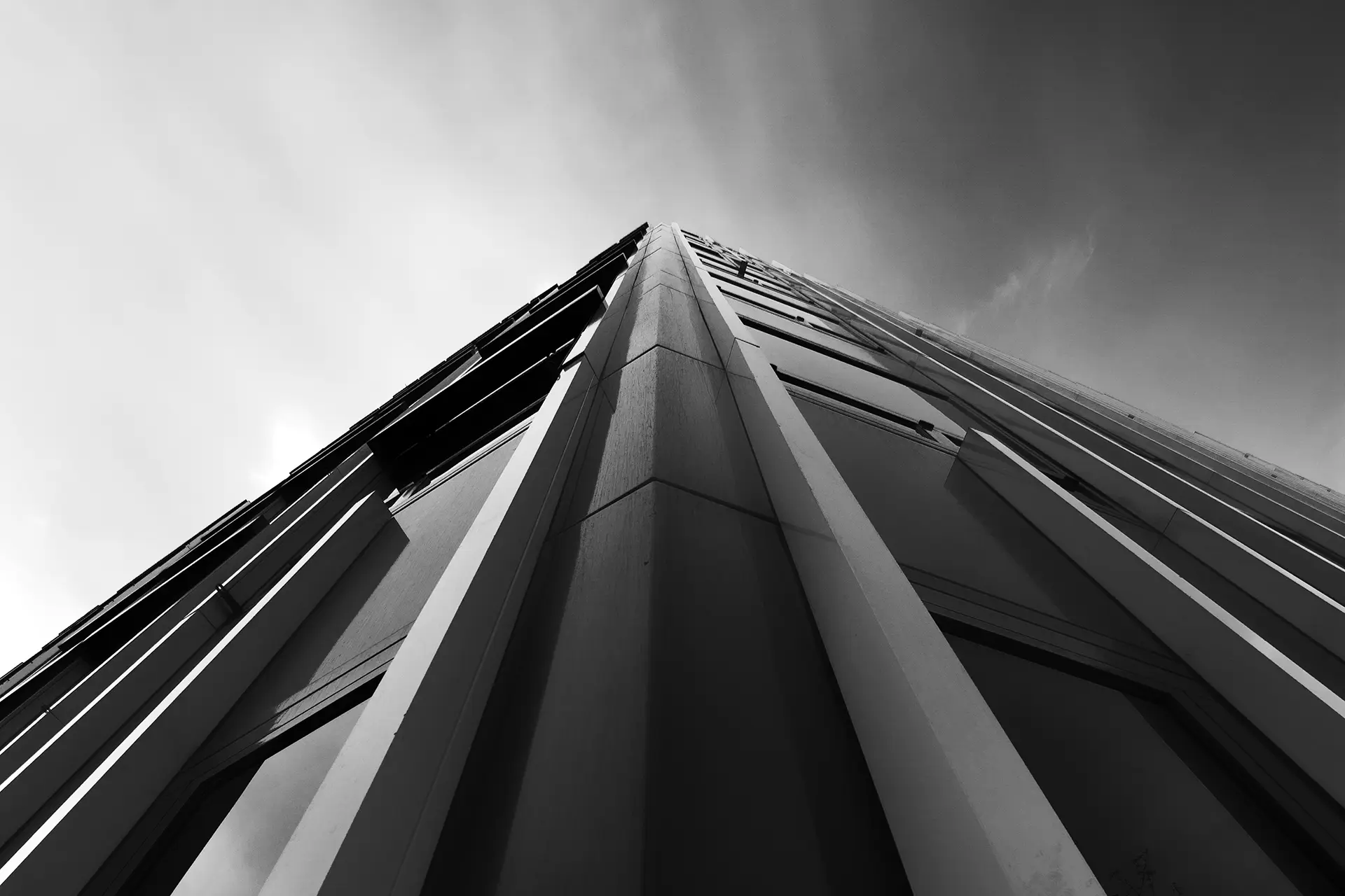 Schwarz-Weiß-Foto: ein Gebäude aus der Nähe am Gebäudeeck mit Blick nach oben aufgenommen. In der Mitte laufen kantige Säulen am Bauwerk nach oben spitz zu in einen dramatischen Himmel.