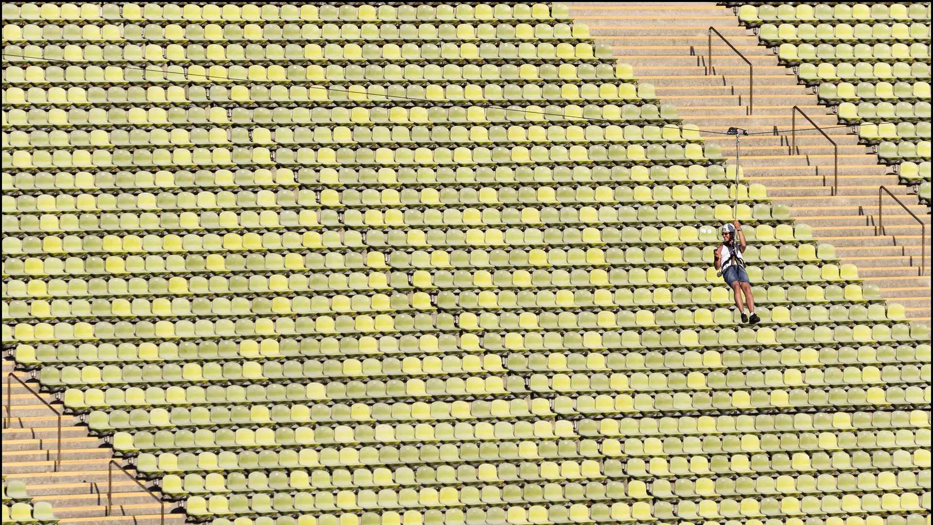 Farbfoto: Privates Event einer Sportlerin, die an einem quer durch das Olympiastadion gespannten Seil an Rollen auf dem Seil von einer Seite des Stadions auf die andere Seite gleitet. Im Hintergrund die leeren grünen Schalensitze.