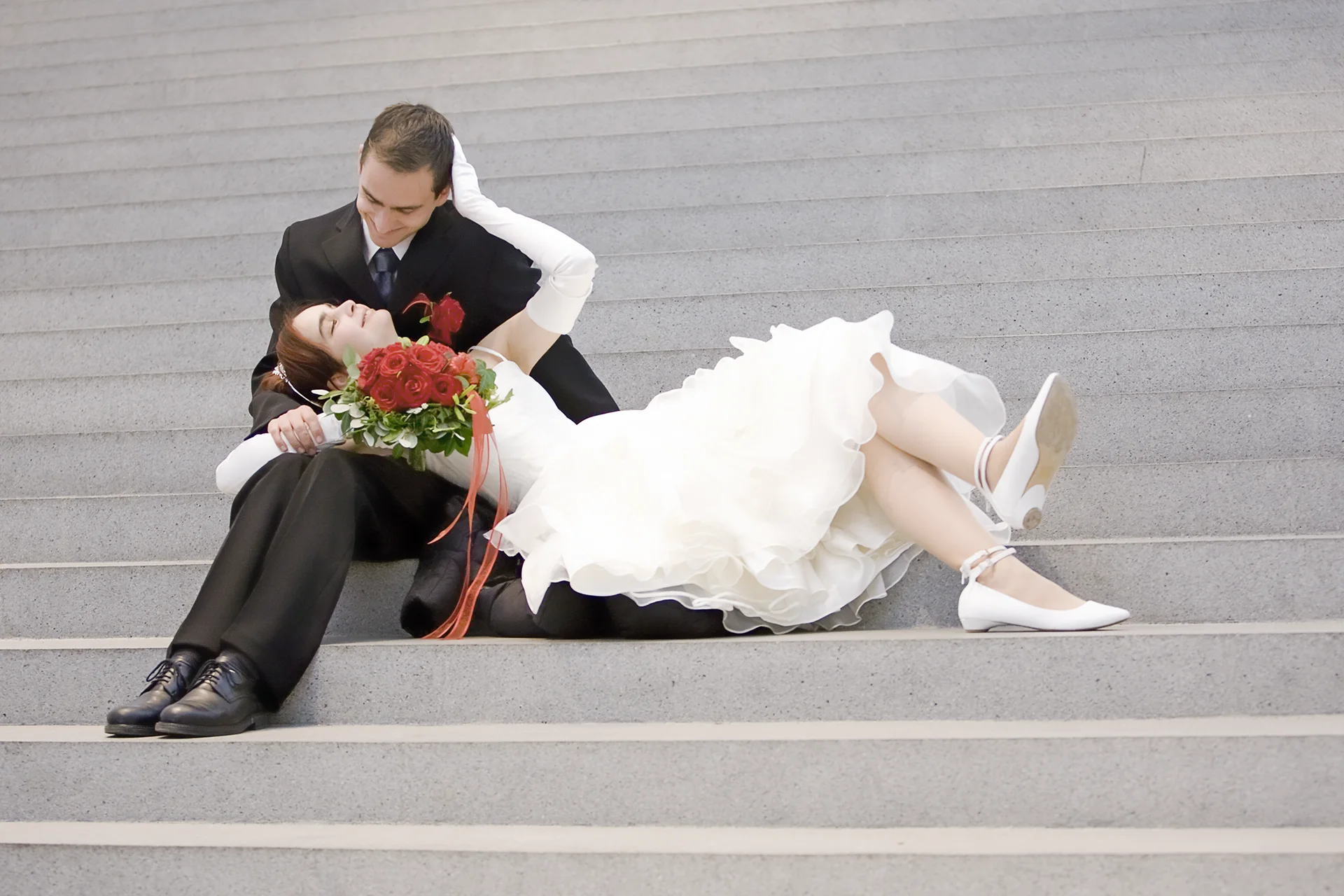 Farbfoto: Ein Brautpaar auf grauen Stufen in einem Gebäude, der Mann sitzt links mit angewinkelten Knien und die Frau im weißen Brautklaid ligt rechts auf den Stufen, den Kopf auf seinen Beinen unmit dem Arm auf seiner Schulter. Die Augen sind aufeinander gerichtet die Köpfe nähern sich an.