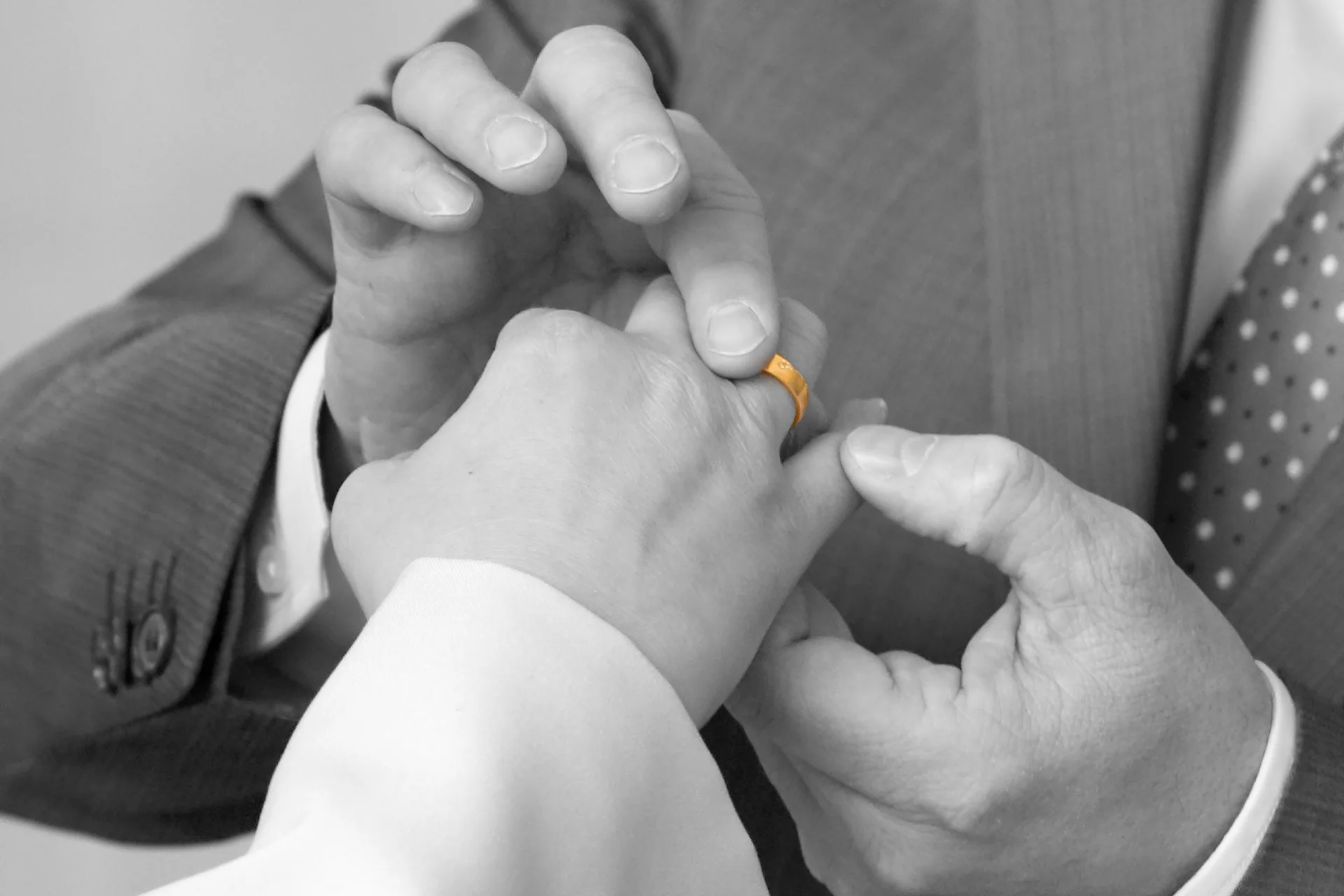 Schwarz-Weiß-Foto mit Ehering in Farbe: Nahaufnahme mit den Händen des Brautpaares, im Moment, als er den goldenen Ring auf ihren Finger steckt.
